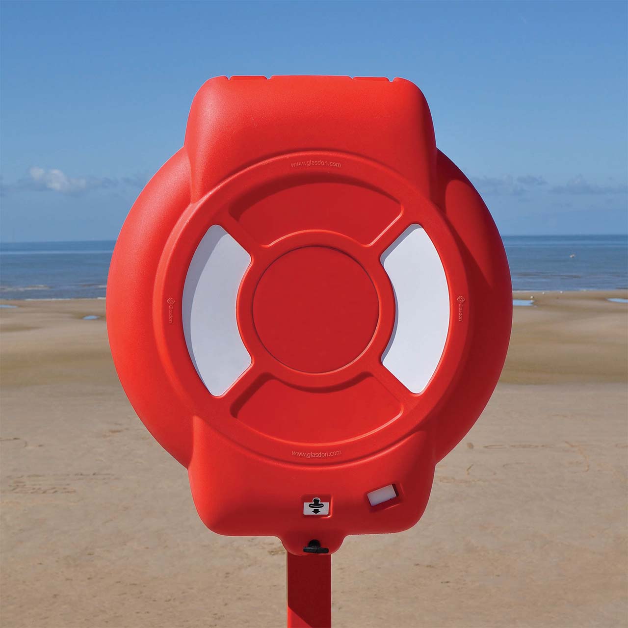 Guardian lifebuoy housing mounted on a post at a beach waterfront, providing highly visible emergency water safety equipment.